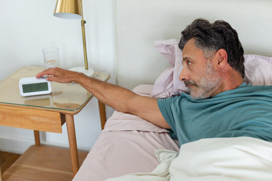 Middle-aged man lying on pink sheets in bedroom reaching toward digital alarm clock in teal shirt