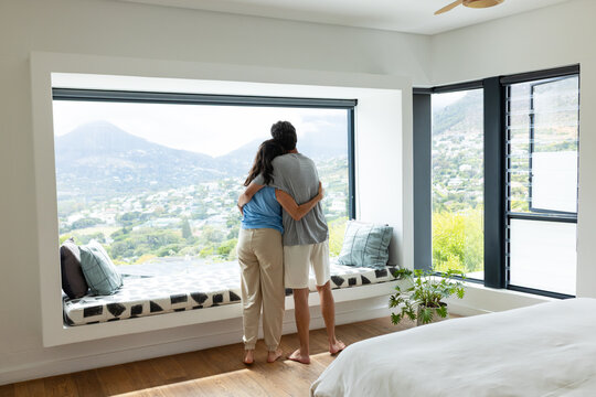 Couple standing on window seat in bedroom facing large panoramic window holding each other