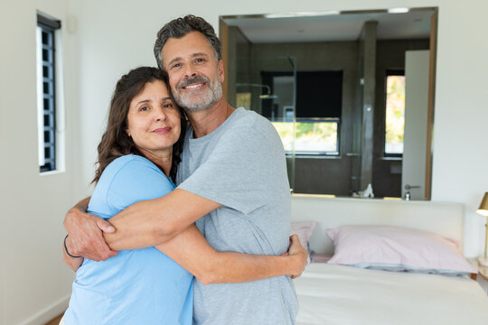 Diverse couple wearing gray and blue t-shirts, hugging, facing camera in bedroom with bed, window