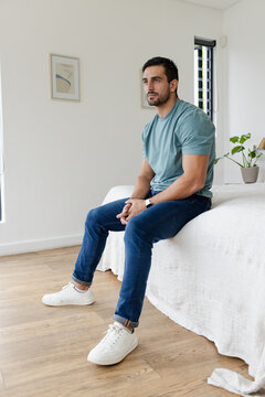 Man sitting on bed edge in simple bedroom with white coverlet, plant, blinds, checking wristwatch