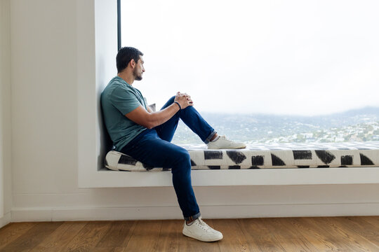 Man sitting on home window bench with bold cushion wearing tee, gazing through window, copy space