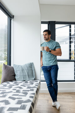 Man leaning against column in bright home, holding beige mug, wearing smartwatch, copy space