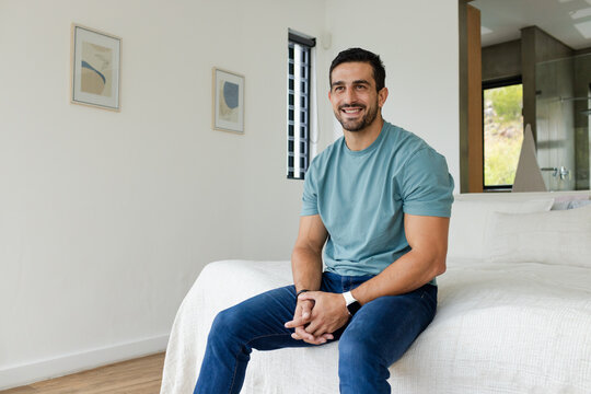 Man sitting on bed edge in bright minimalist bedroom wearing teal shirt and wristwatch and bracelet
