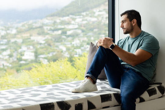 Man sitting on patterned bench at home, gazing through rainy window, wearing smartwatch, copy space