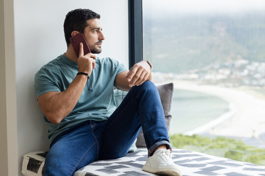 Man sitting on window seat wearing teal tee, holding maroon phone with smartwatch, gazing at coast