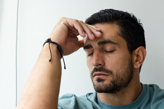 Adult male seated in room, closing eyes, touching brow left-hand after work, teal-shirt black-cord