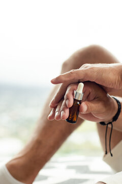 Adult male holding amber dropper bottle, preparing to apply serum onto palm beside bright window
