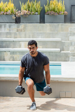 Adult male lowering into weighted lunge at poolside holding hex dumbbells, wearing dark gray shirt