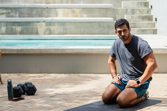 Man late 20s kneeling on mat at poolside, wearing shorts, smartwatch and dumbbells, copy space