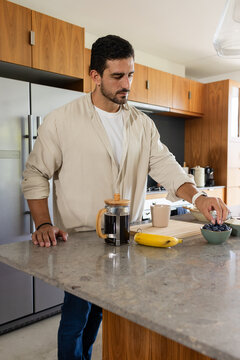 Man standing at kitchen island reaching for bowl of blueberries beside French press coffee