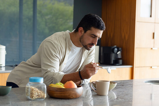 Man wearing light shirt leaning over stone island in kitchen, scooping granola with spoon into mug