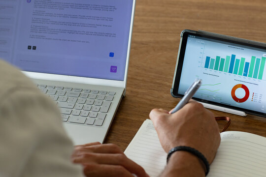 Adult male writing in notebook with pen at wooden desk while referencing laptop, tablet charts