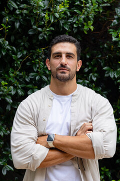 Man standing by hedge with arms crossed, facing camera wearing beige shirt, white tee, smartwatch
