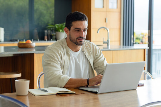Man working on silver laptop at wooden dining table in modern kitchen, notebook, ceramic mug nearby