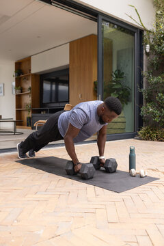African man wearing athletic clothes holding hex dumbbells, performing plank on mat on brick patio