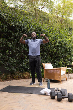 African American adult man flexing arms in athletic wear on backyard patio near mat, dumbbells