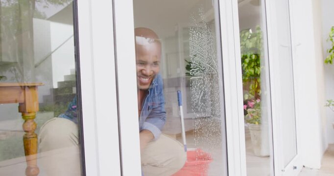 Adult African American man in denim noticing soapy film, cleaning sliding-door entry with squeegee