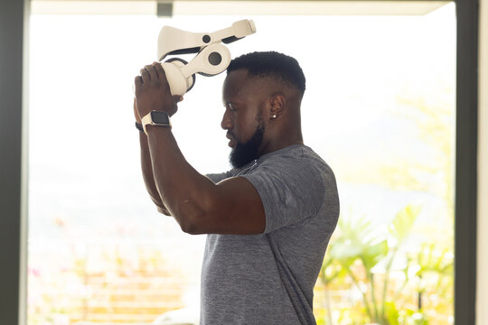 African American man holding white VR headset above head, wearing grey shirt near glass doors