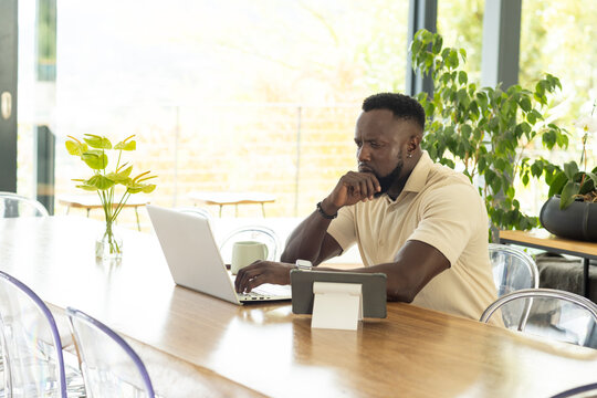 African man sitting at table, wearing beige polo, typing on laptop, thinking on report, copy space