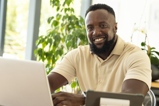 Man wearing beige polo sitting at desk using laptop with tablet stand, potted plants by windows
