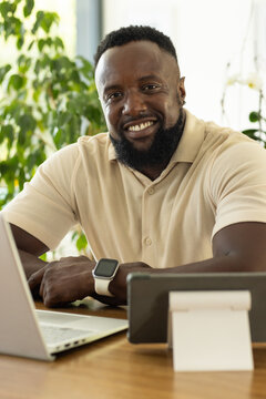 African man sitting at oak desk in sunlit plant office wearing polo and smartwatch using laptop-tab