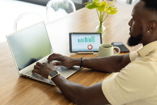 African American adult man typing at wooden table on laptop with tablet, mug, phone, copy space
