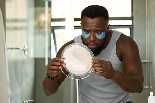 African American man inspecting face with magnifier at sink wearing blue under-eye patches