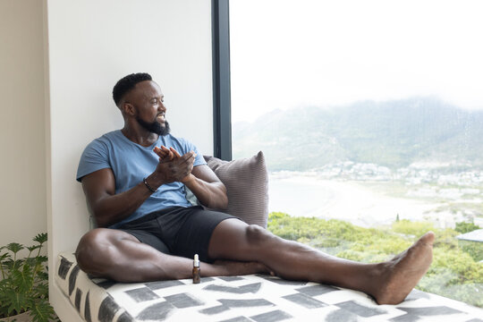 African man sitting on window bench in sportswear, rubbing hands near dropper, copy space