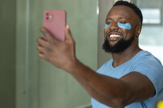 African man taking selfie with pink phone while wearing blue eye patches in bathroom