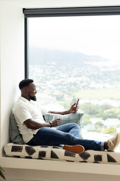 Adult African man taking break, holding mug and scrolling smartphone on window bench, copy space