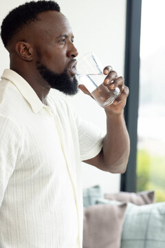 African man drinking from clear glass in living room near window wearing cream textured shirt