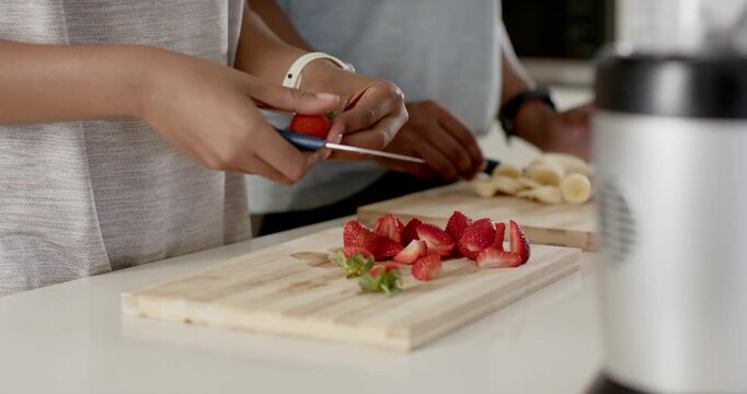 Couple slicing strawberries and bananas at kitchen counter with knives, preparing fruit for blender