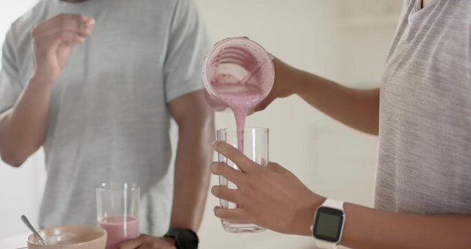African American couple woman tipping jar, pouring pink smoothie into glass in kitchen to drink