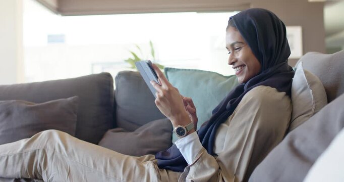 Woman in hijab reclining on sofa with wristwatch, glancing at camera then tapping tablet to read