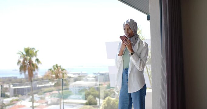 Adult woman in headscarf standing on balcony after message alert, scrolling pink phone, copy space
