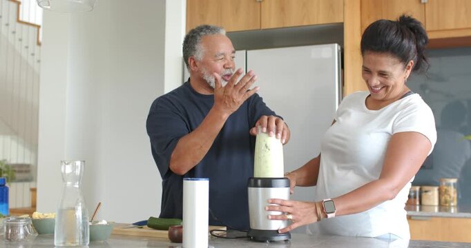 Senior African American couple placing blender jar with green mixture, making smoothie on island