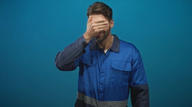 Man with beard in blue work overalls covers face with hand while smiling and peeking through fingers in studio; embarrassment.