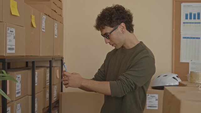 Man checking smartphone with hands while scanning parcels on shelves in a building; concentration productivity.