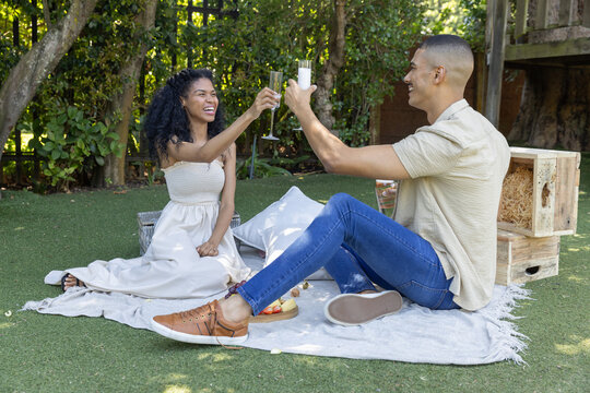 Couple in white strapless dress and jeans clinking champagne flutes on picnic blanket in backyard