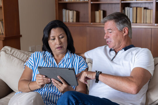Diverse couple sitting on beige sofa in living room, holding black tablet and pointing at bookshelf