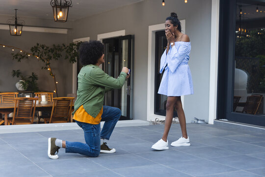 African American couple kneeling on patio, proposer holding open ring box, string lights glowing