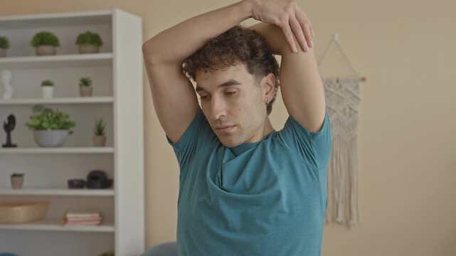 Man stretches arm over head in a studio with shelf and plants visible indoors; serenity focus mindfulness.