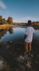 Fototapeta premium Man standing near a river observing the flowing water, surrounded by trees in a peaceful natural landscape