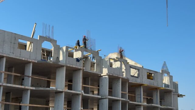 Builders on rooftop installing concrete slabs under clear blue sky, crew in helmets guiding crane and positioning precast panels.
