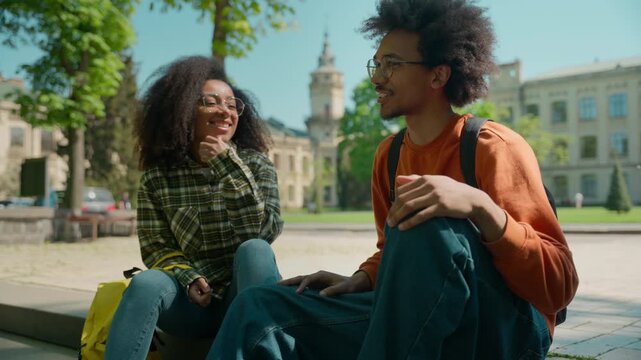 Two African American students talking together smiling sharing news outdoors couple man guy male boyfriend conversation laughing woman female girl girlfriend friends communicate campus city university
