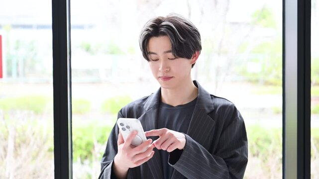 新緑の窓際でスマートフォンを操作する検索する若い男性社員のビジネスと日常のシーンのクローズアップの動画	