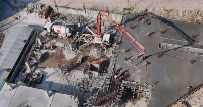 aerial view of a bustling construction site featuring an orange concrete pump truck and workers casting long shadows as they level fresh cement over an intricate rebar grid.