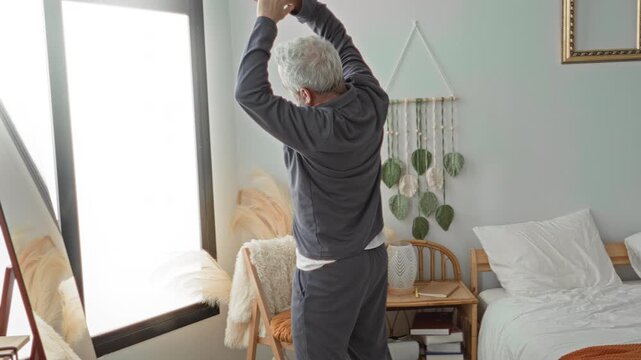 Middle age man with grey hair stretches hands above head beside bed in a bright bedroom inside a building, facing a mirror and woven wall hanging; serenity.