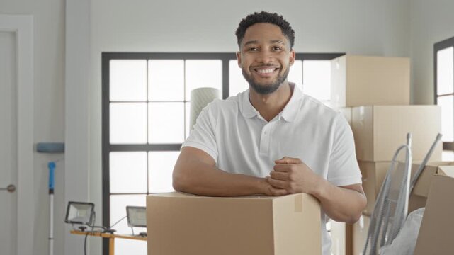 Smiling man holds a cardboard box, rests arm on it and raises fist beside stacked boxes and floodlight in building; moving day confidence.