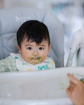 Happy Asian baby boy in high chair smiling at mother while being spoon-fed. Close-up view with natural light in a home setting.	
